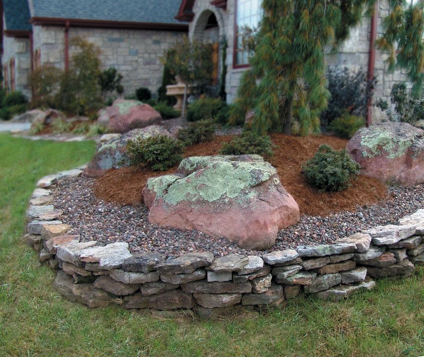 A stone-walled garden bed with large lichen-covered boulders, mulch, and small shrubs in front of a stone house.