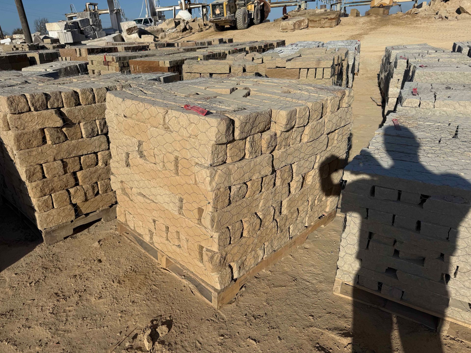 Pallets of tan stone building blocks stacked outdoors in a construction or supply yard under a bright, sunny sky.
