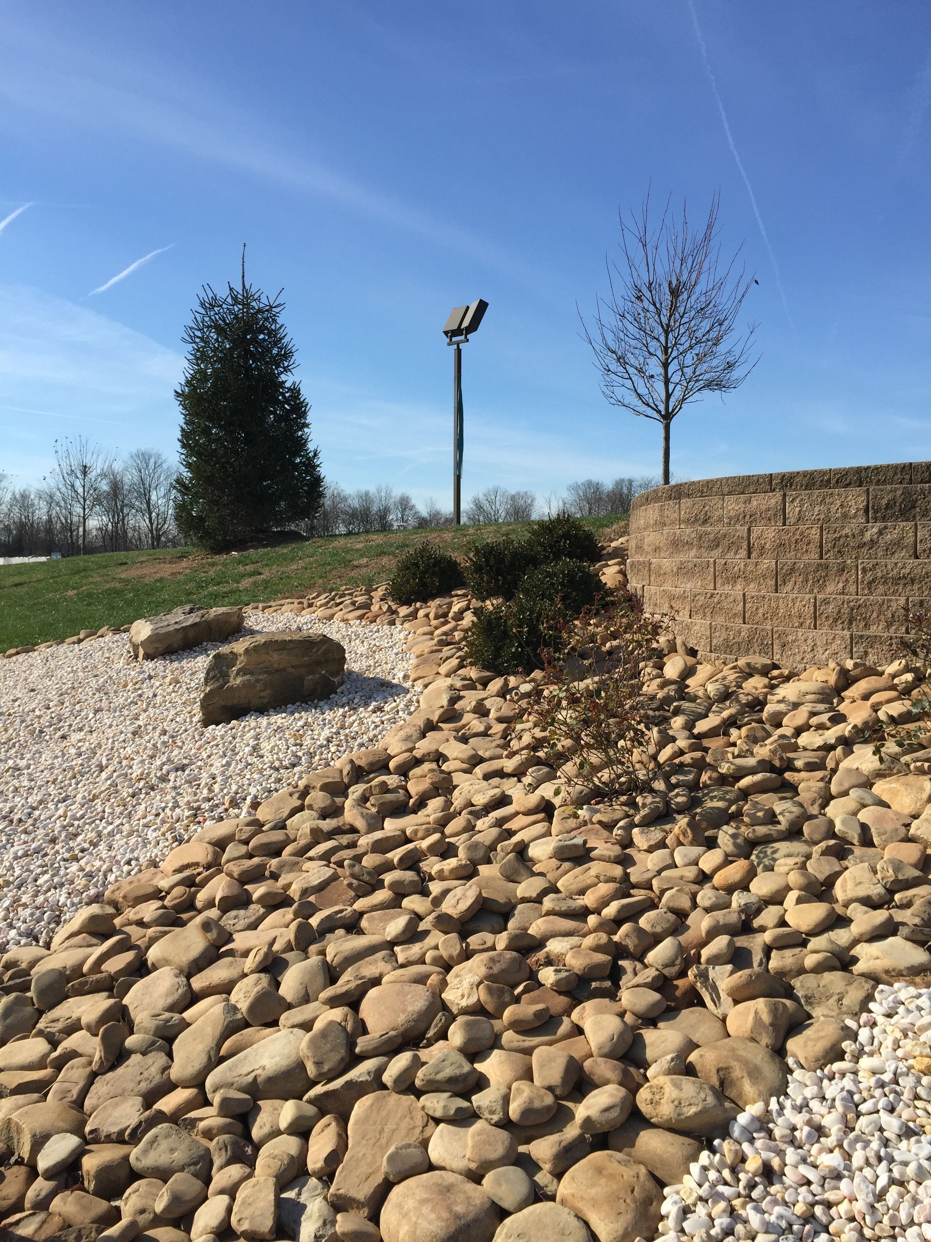 A landscaped garden featuring a stone retaining wall, tan river rocks, and white gravel under a clear blue sky.