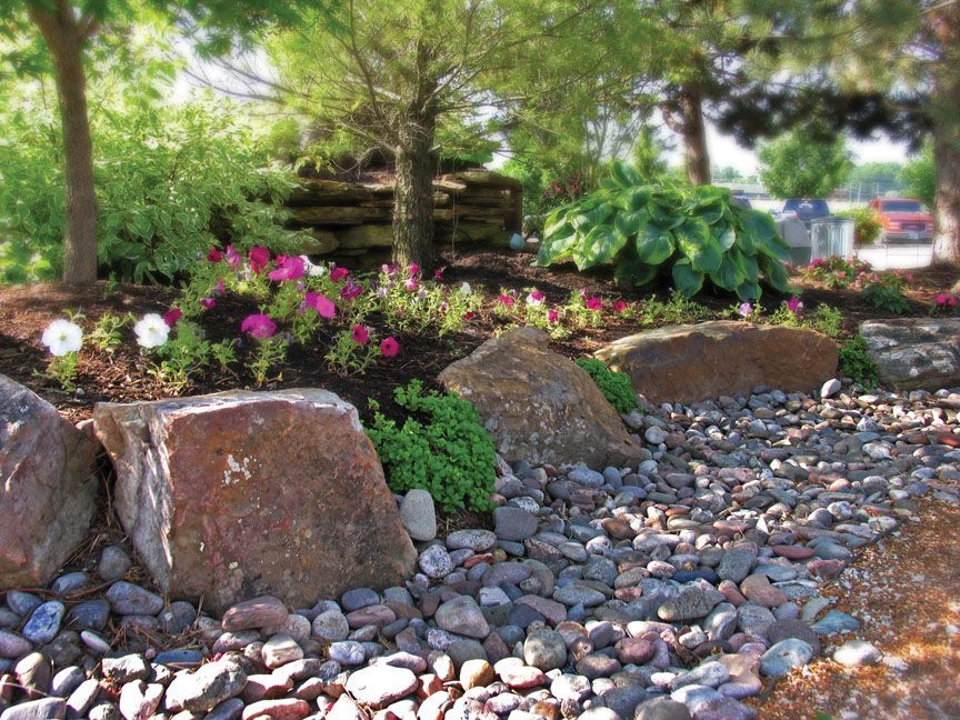 A landscaped garden bed with large brown boulders, dark mulch, pink flowers, green foliage, and a bed of river stones.