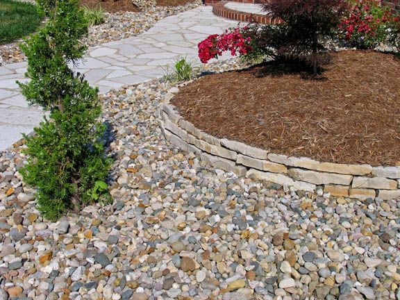 A landscaped garden bed with mulch and a stone border, surrounded by decorative pebbles and a spiral-shaped evergreen.