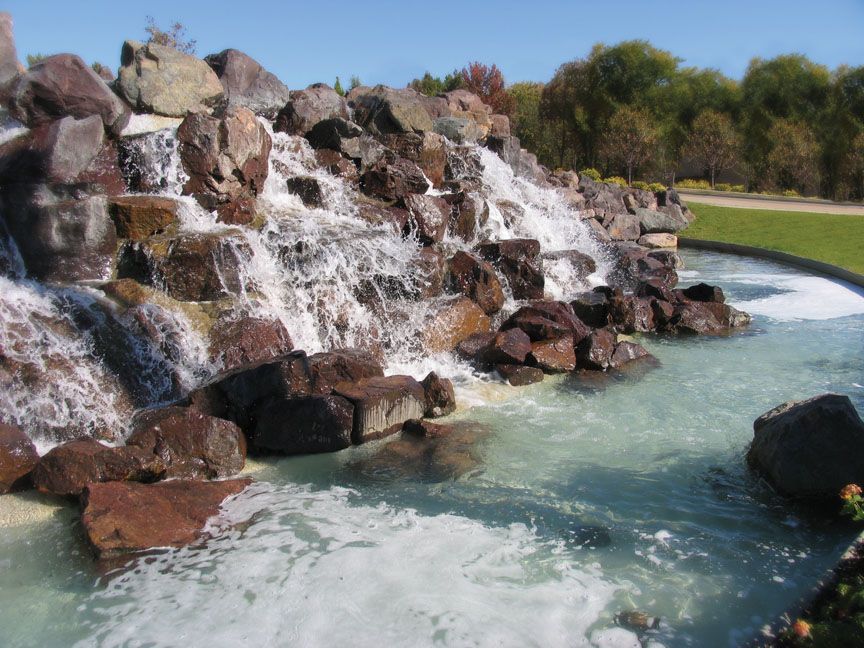 A rocky, man-made waterfall flows into a turquoise basin, surrounded by green grass and trees under a clear blue sky.