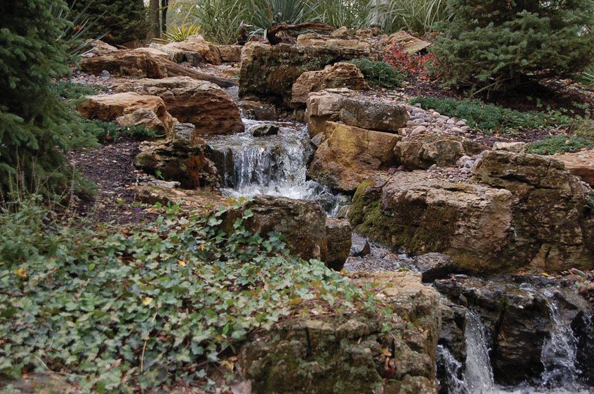 A rocky garden waterfall cascading over tan stone blocks, surrounded by greenery and ivy.