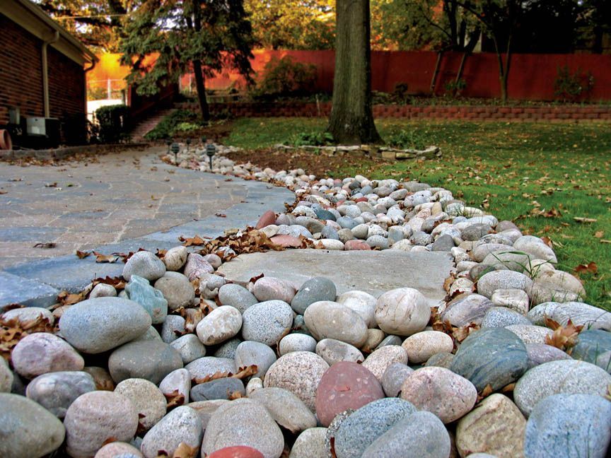 A curved border of smooth river rocks separates a paved patio from a green lawn and a large tree in a backyard setting.