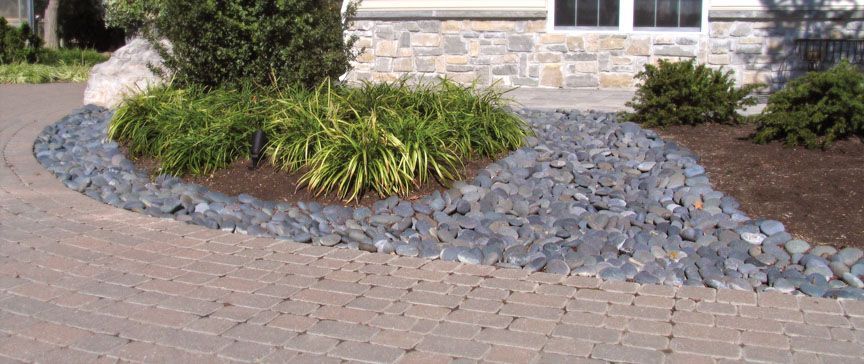 A curved stone paver walkway bordering a garden bed with green shrubs, dark mulch, and a decorative gray rock border.