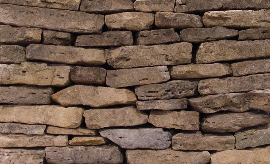 A close-up view of a rustic, dry-stacked stone wall with irregular, weathered tan and brown stones.