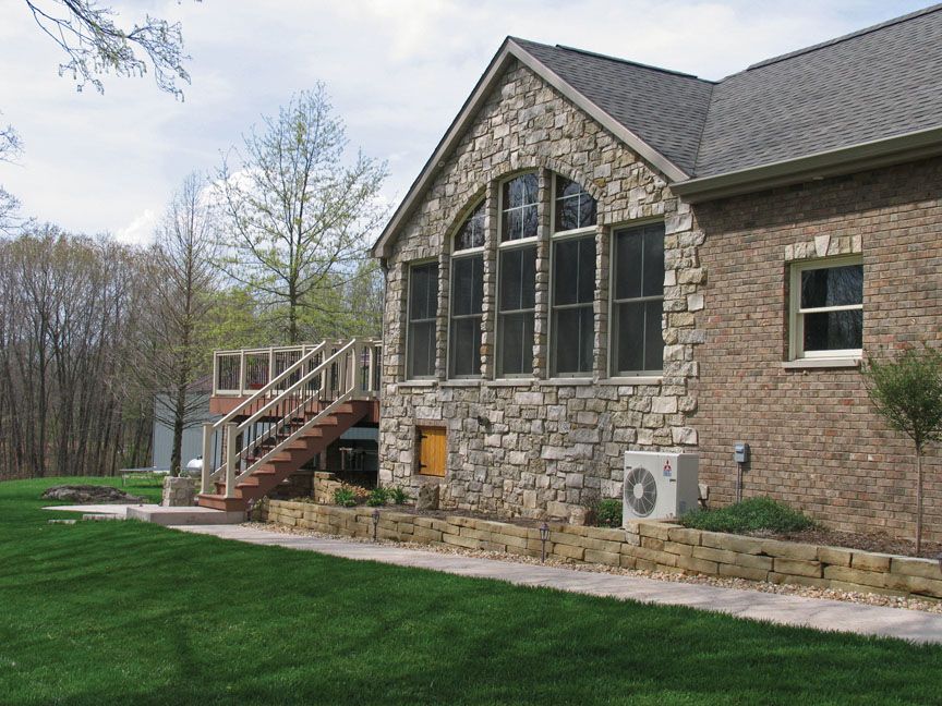 A stone house with an arched window, a wooden deck with stairs, and a stone retaining wall in a grassy backyard.