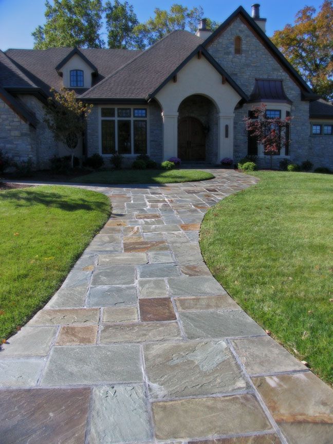 A stone walkway leads to the entrance of a traditional home with stone siding and a gabled roof on a sunny day.
