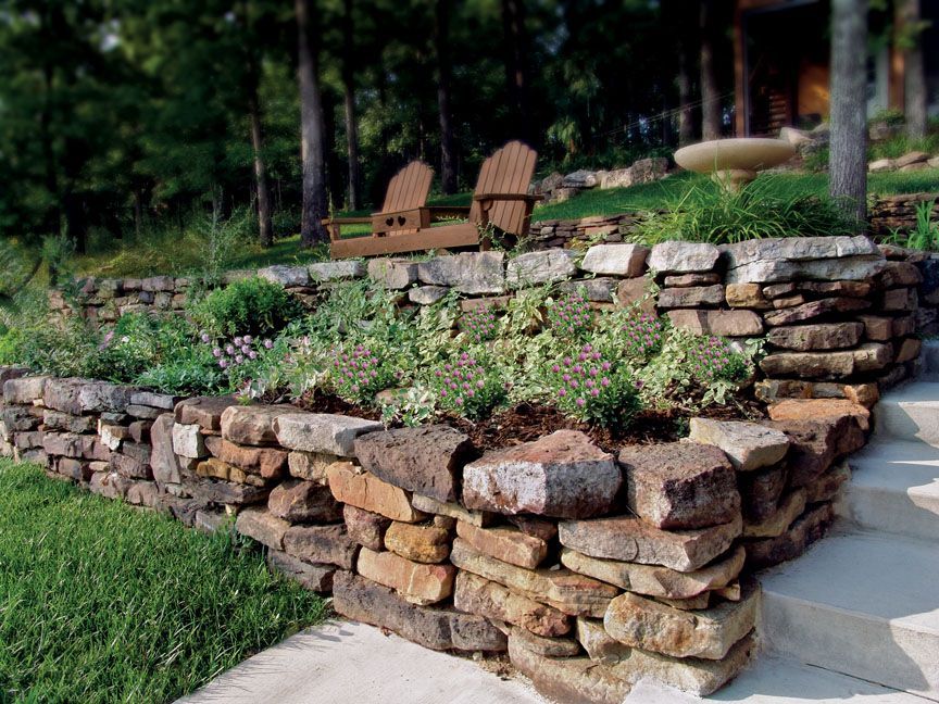 A stone retaining wall filled with lush green plants sits beside concrete steps leading up to wooden patio chairs.