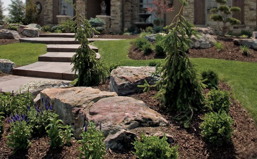 A flagstone walkway leads to a stone house through a landscaped garden with boulders, conifers, and shrubs.