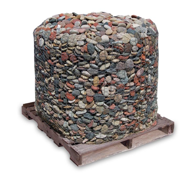 A large, cylindrical wire cage filled with multicolored decorative river stones sitting on a wooden shipping pallet.