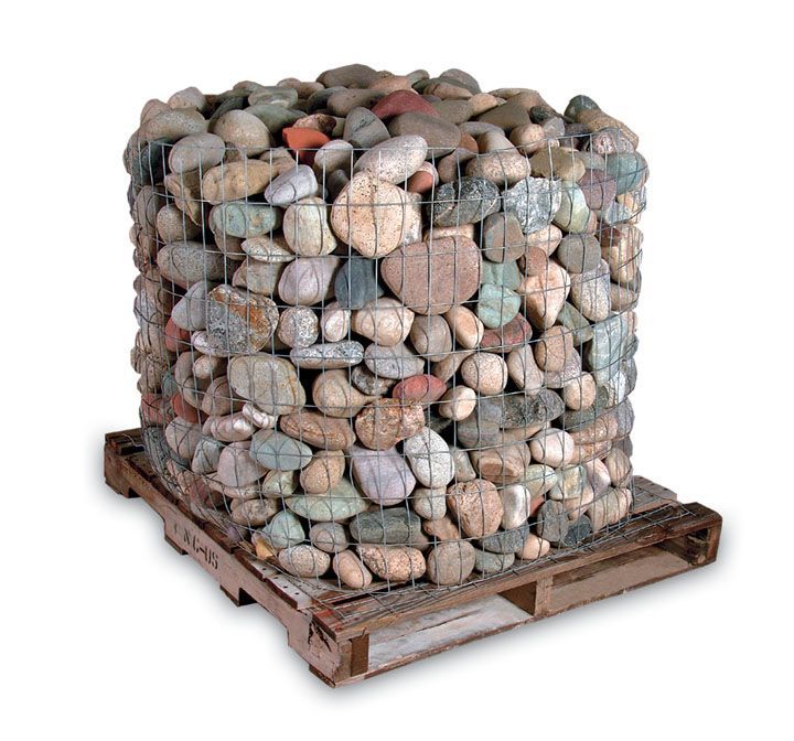 A wire cage filled with smooth, multicolored river rocks, sitting on a wooden shipping pallet against a white background.