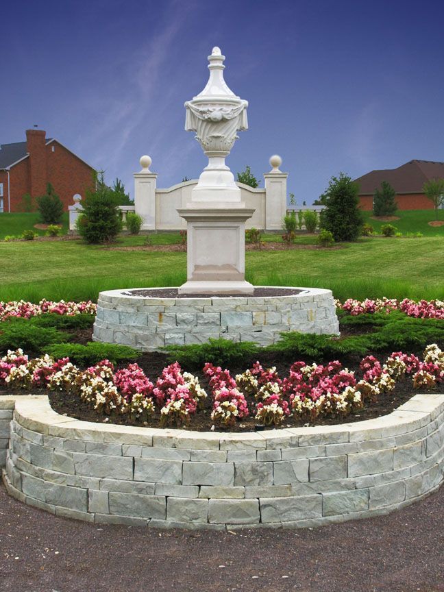 A stone pedestal with a decorative urn centerpiece, surrounded by flowers and greenery in a residential neighborhood.