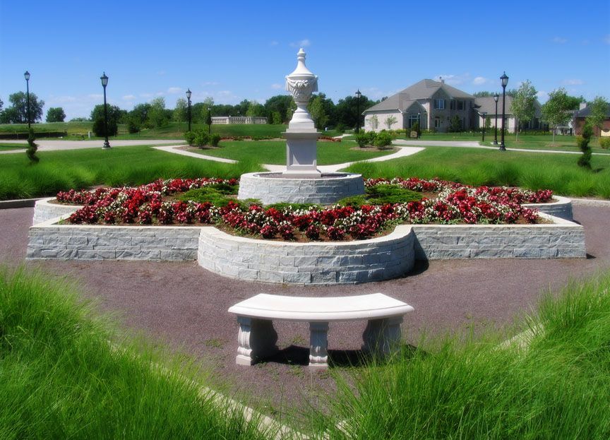 A stone fountain with red flowers in a star-shaped bed, featuring a curved bench in front against a suburban backdrop.