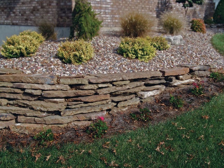 A tiered landscape bed with a natural stone retaining wall, mulch, shrubs, and a brick house in the background.