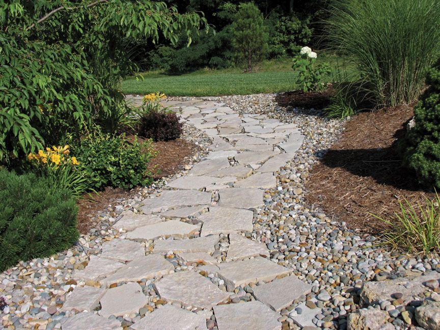 A light gray flagstone garden path winds through a landscape with small shrubs, yellow flowers, and gravel borders.