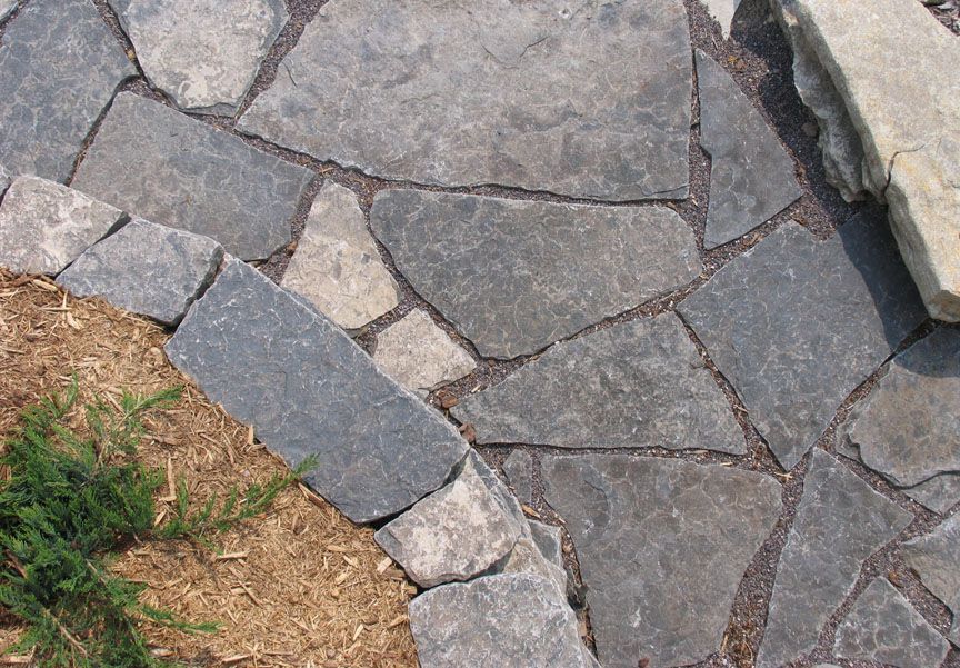 A close-up of a flagstone walkway with dark gray, irregular stones edged by a curved row of pavers and mulch.