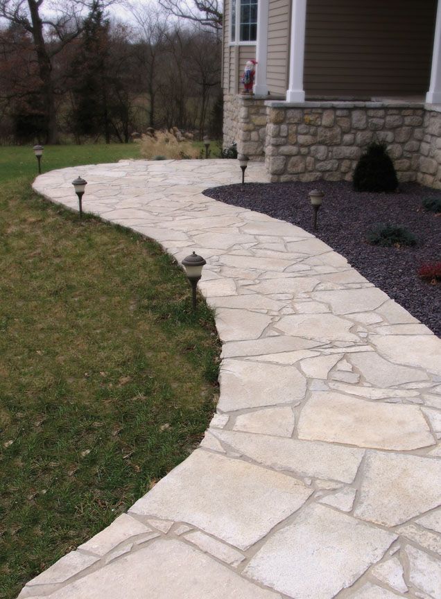 A curved flagstone walkway leads toward the entrance of a house with stone siding and a lawn to the side.