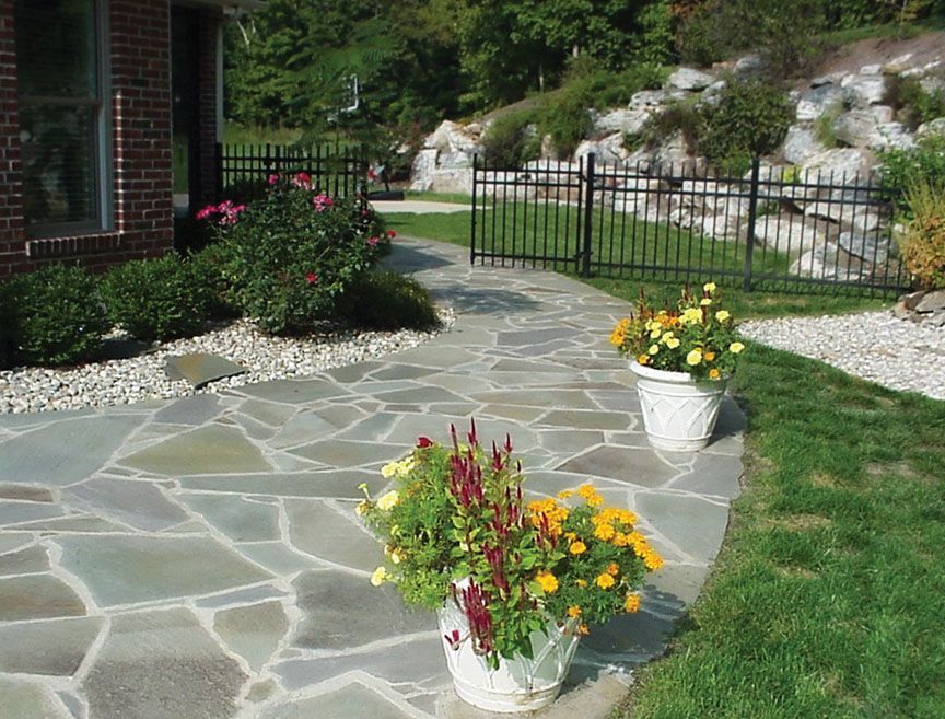 A stone patio walkway curves toward a black metal fence, lined with white planters filled with colorful flowers.