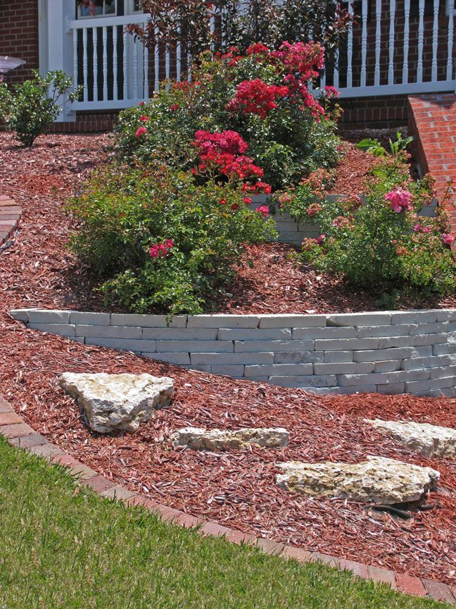 A tiered garden bed with red roses, tan stone retaining walls, red mulch, decorative rocks, and a brick-edged lawn.