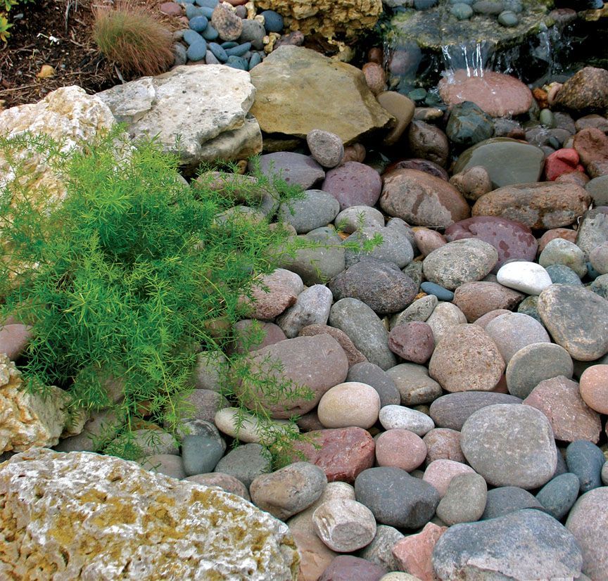 A decorative landscape feature with a small waterfall flowing over stones, colorful river rocks, and lush green plants.