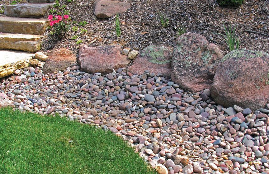 A garden bed features a border of large, weathered granite boulders, river rock mulch, and stone steps beside green grass.