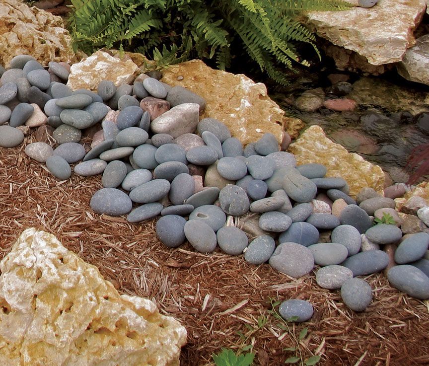 A close-up of smooth, gray river stones bordering mulch next to large, textured tan landscaping rocks and green plants.