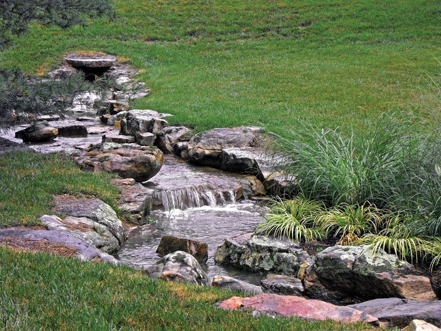 A small, rocky artificial stream with a gentle waterfall flowing through a grassy green lawn.