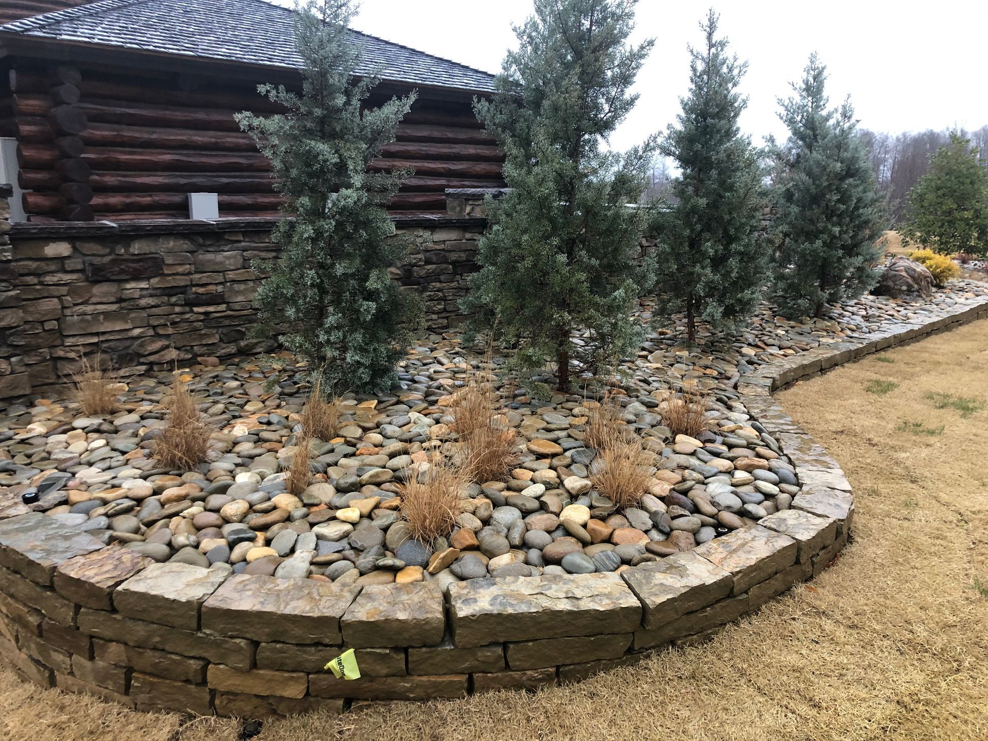 A curved, stone-retaining wall borders a garden bed filled with river rocks, small brown shrubs, and several pine trees.