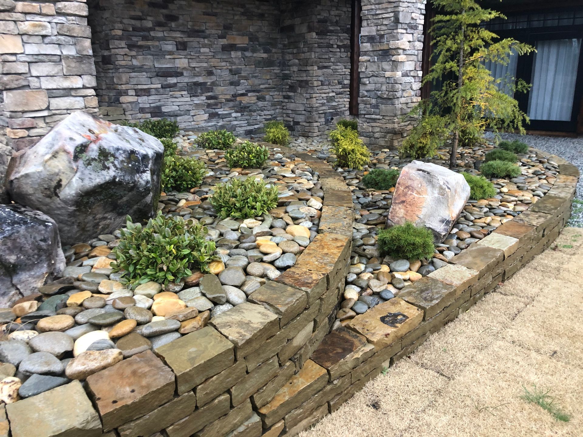 A curved stone retaining wall borders a garden bed filled with river rocks, small green shrubs, and two large boulders.