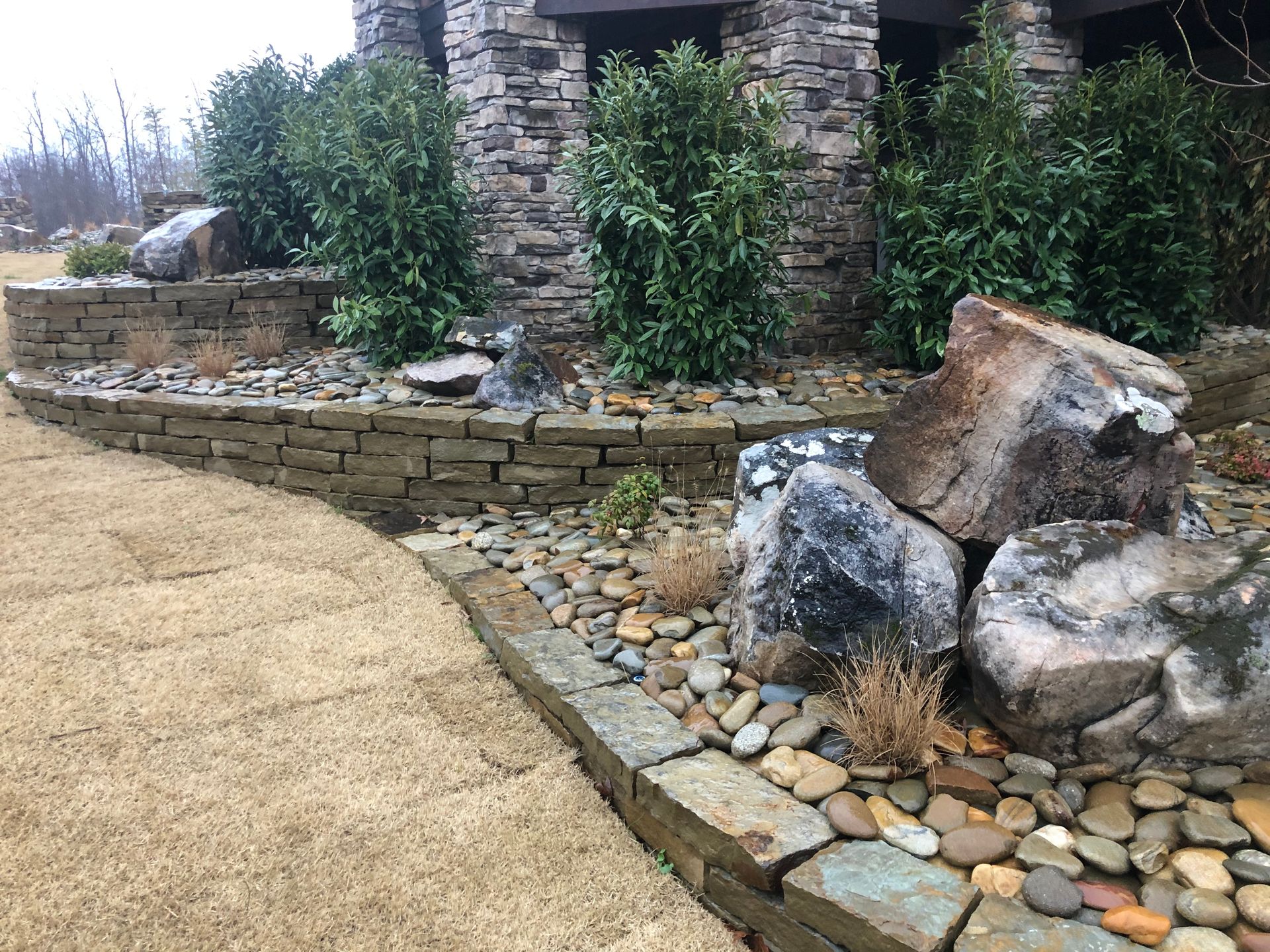 A curved stone retaining wall borders a tan walkway, featuring river rocks, large boulders, and lush green shrubs.