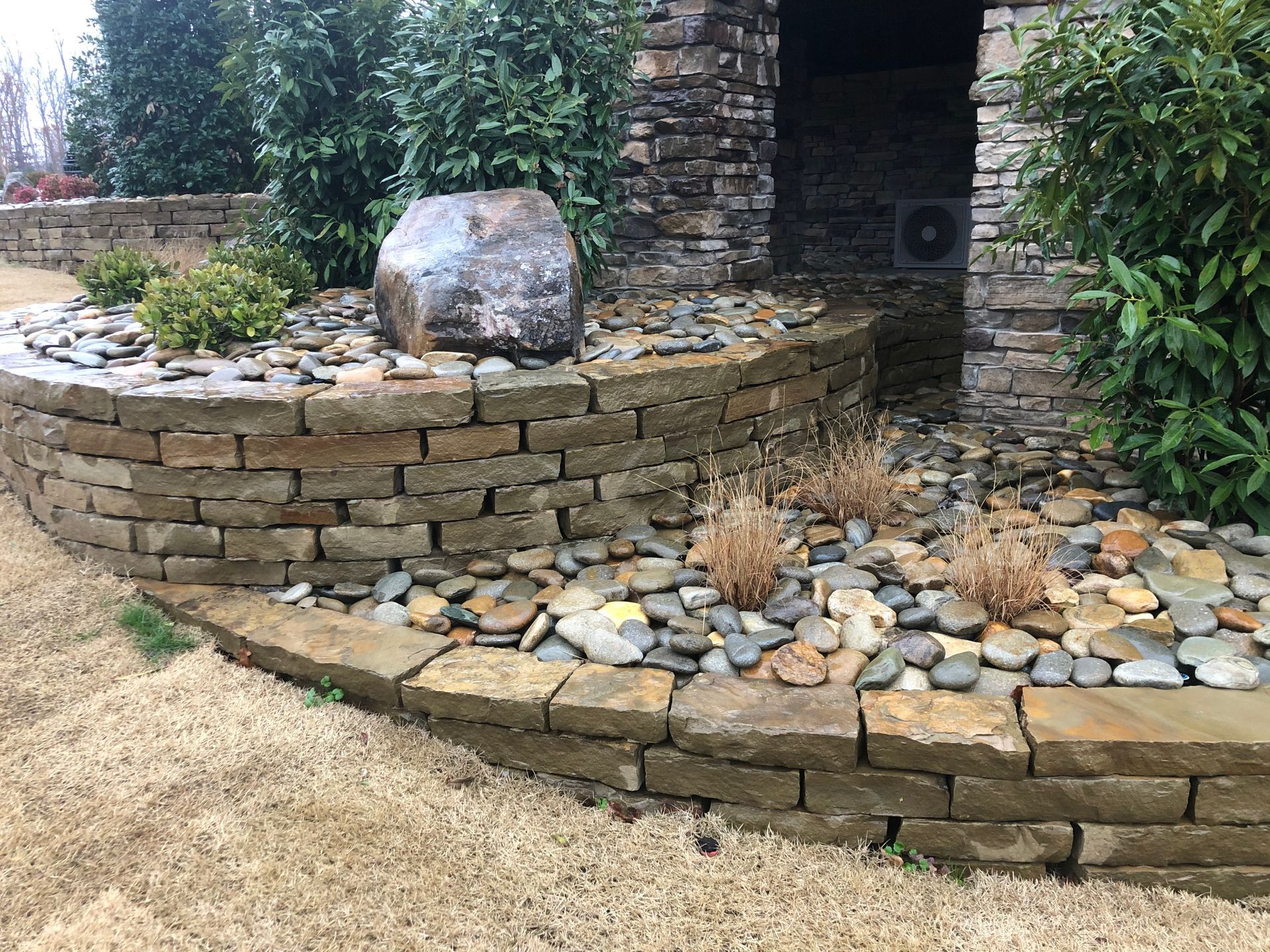 A tiered stone retaining wall with decorative rocks, small shrubs, and a large boulder in a landscaped yard.