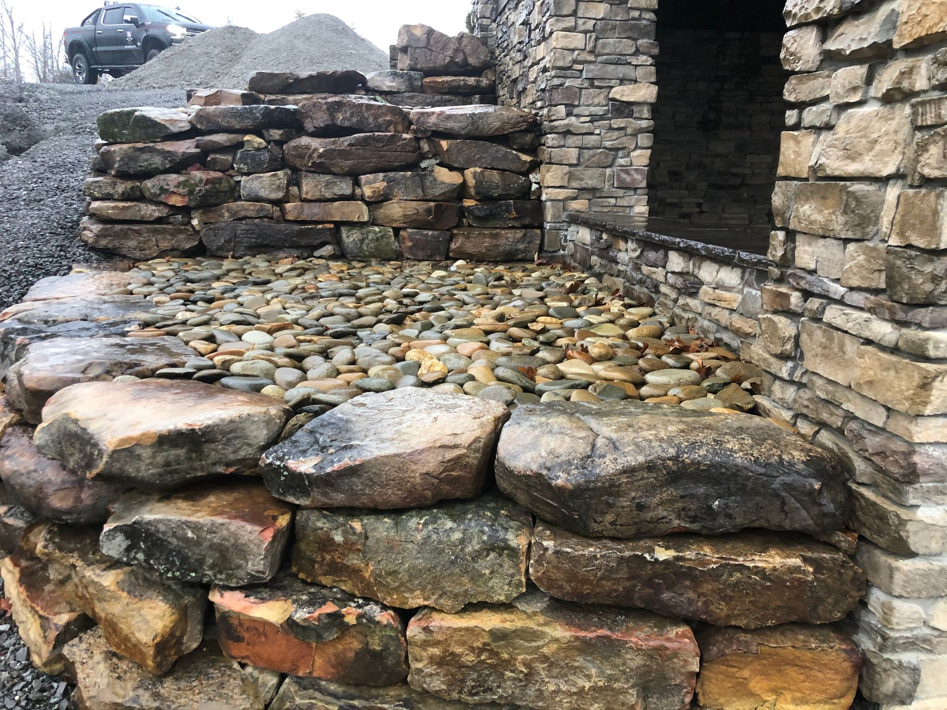 A stone patio with a rounded edge, built with large boulders and smaller river stones, next to a stone building wall.