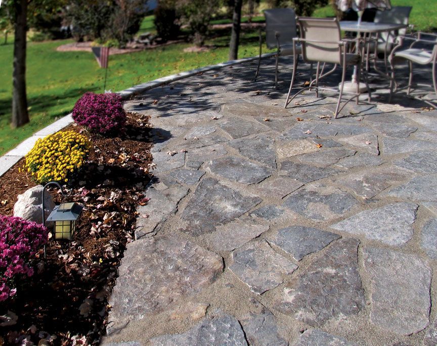 A stone patio with a dining set and a garden bed planted with purple and yellow mums.