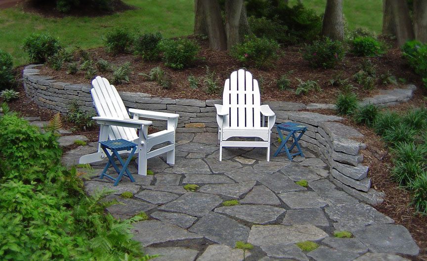 Two white Adirondack chairs with small blue stools on a gray stone patio set against a curved stone retaining wall.