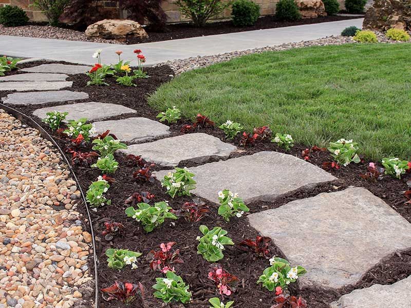 A stone path winds through a garden bed planted with white flowers and dark foliage, adjacent to grass and gravel.
