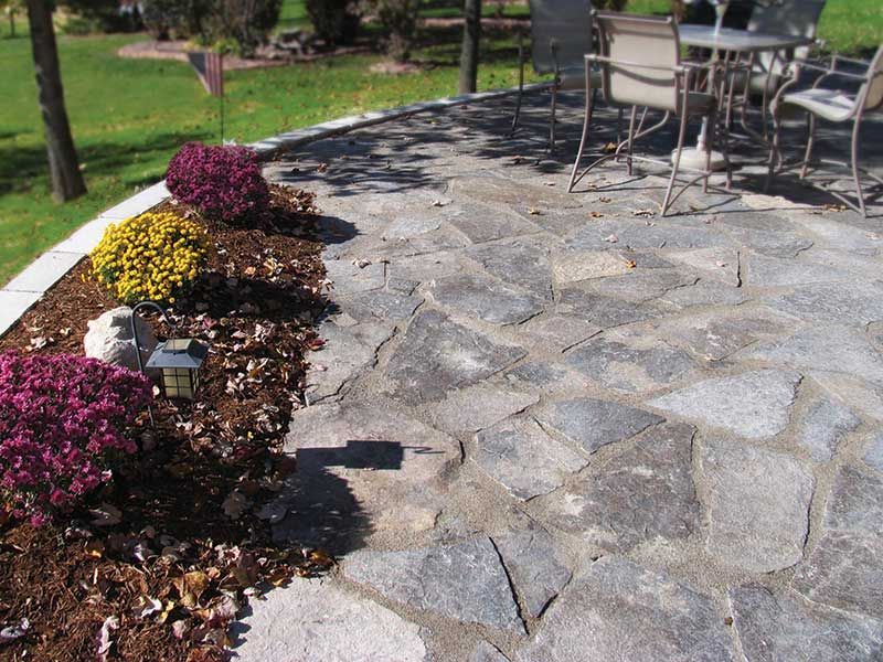 A flagstone patio with a curved flower bed containing colorful mums, next to an outdoor table and chair set on grass.