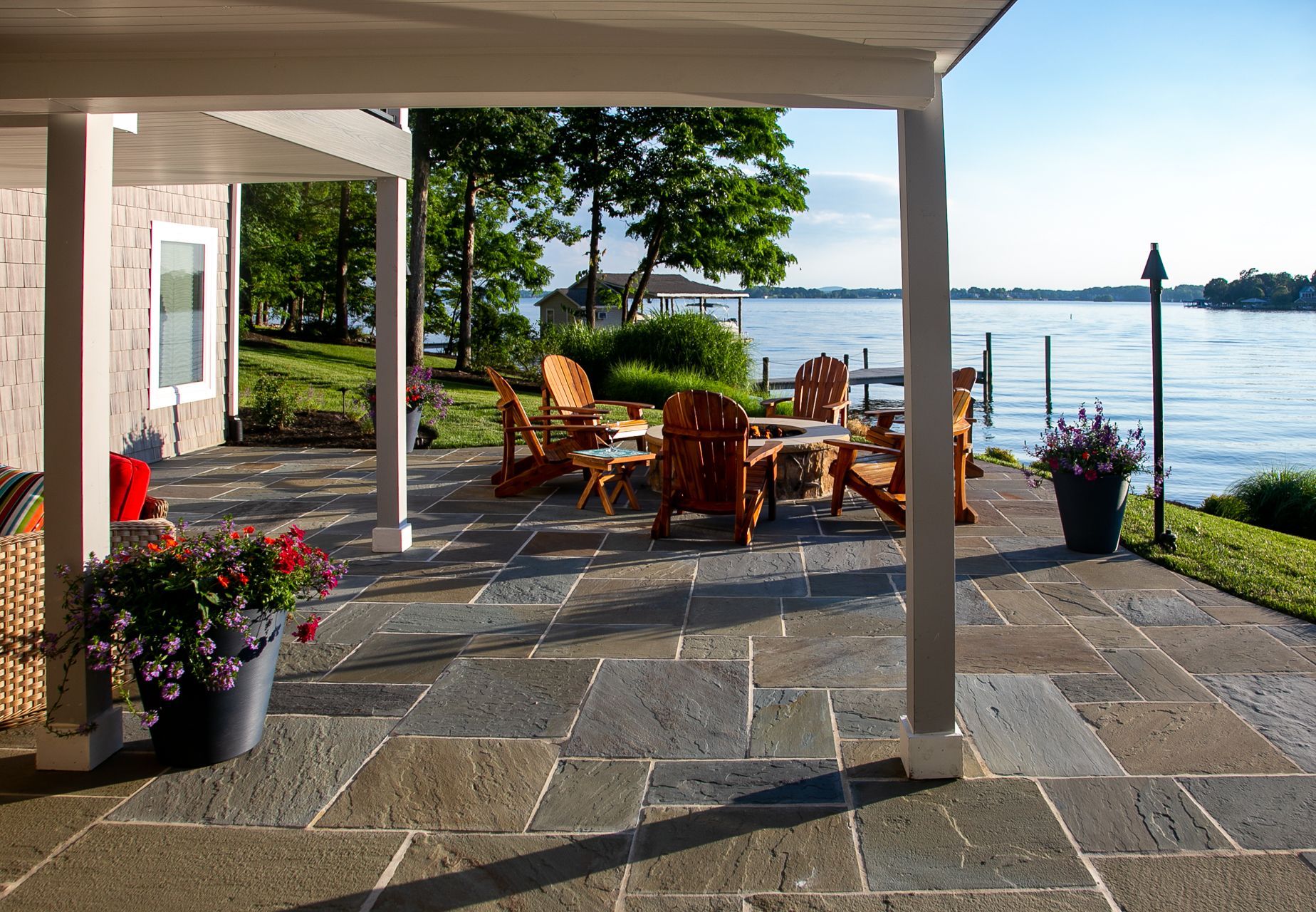 A stone patio with Adirondack chairs and a fire pit overlooks a tranquil lake on a sunny day.