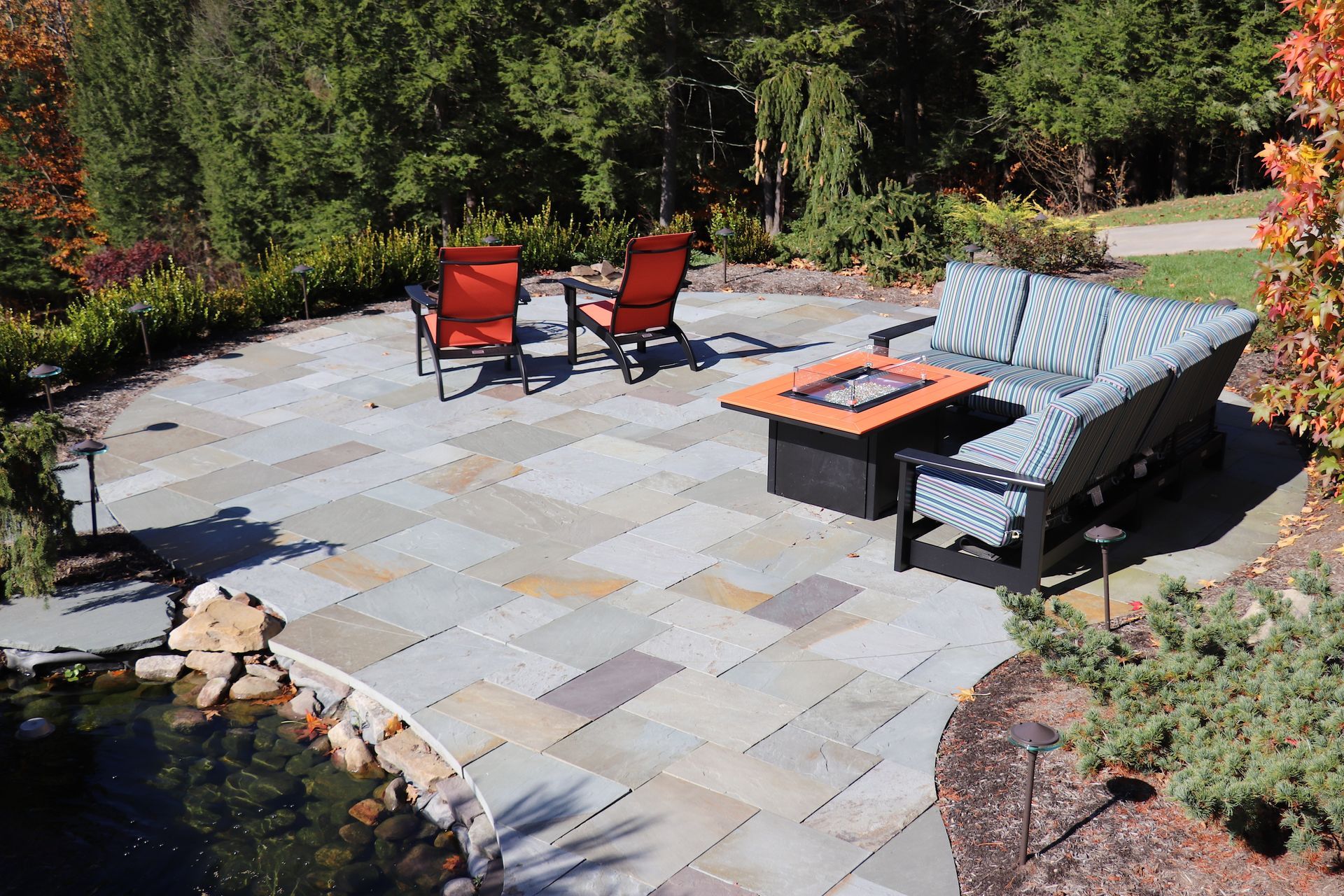 A stone patio with a fire pit, cushioned sofa, and two orange chairs next to a small garden pond in a wooded setting.