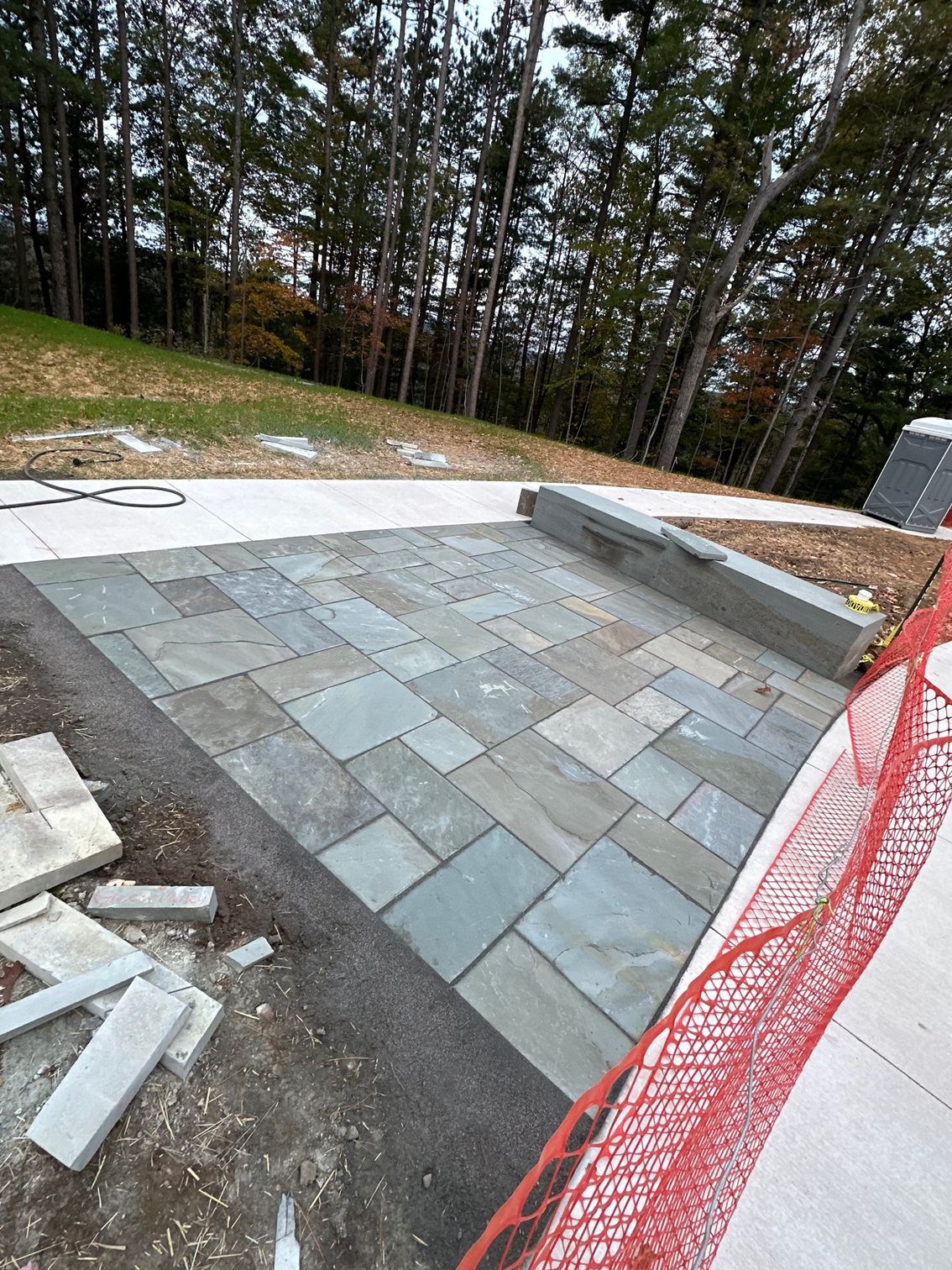 A blue-grey stone paver patio under construction, with a concrete bench and a light-colored sidewalk, near a treeline.