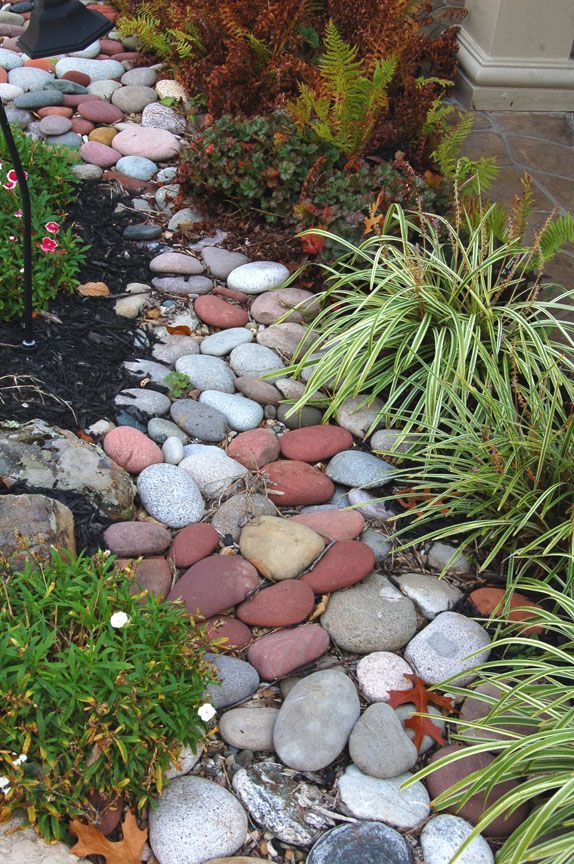A landscaping border filled with smooth, multicolored river rocks, flanked by green bushes and decorative grasses.