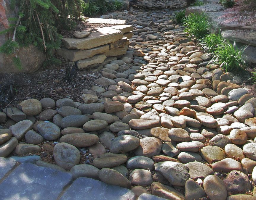 A stone-lined dry creek bed with large boulders, stacked stones, and small green plants in a garden landscape.