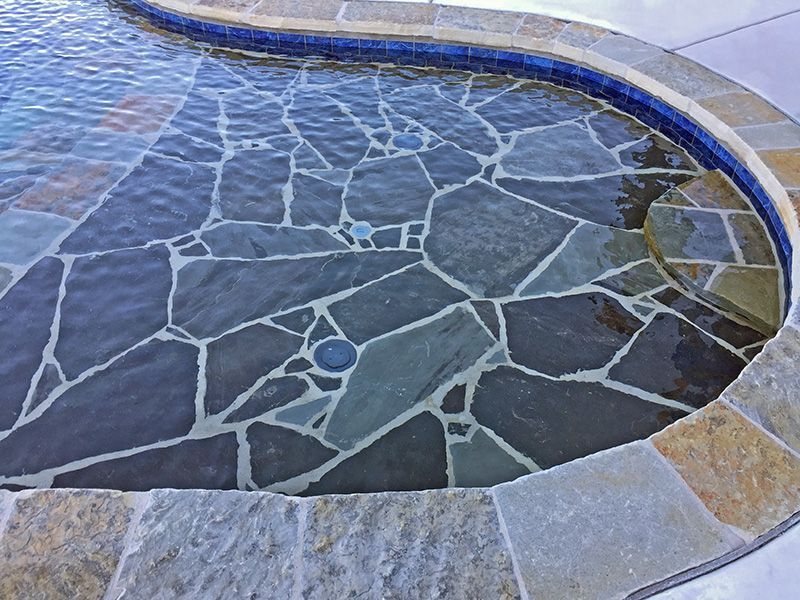 A submerged flagstone bench inside a swimming pool, bordered by blue mosaic tiles and light-colored stone coping.