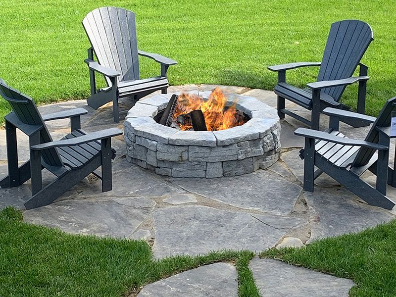 Four gray Adirondack chairs arranged around a circular stone fire pit with a burning fire in a grassy backyard.