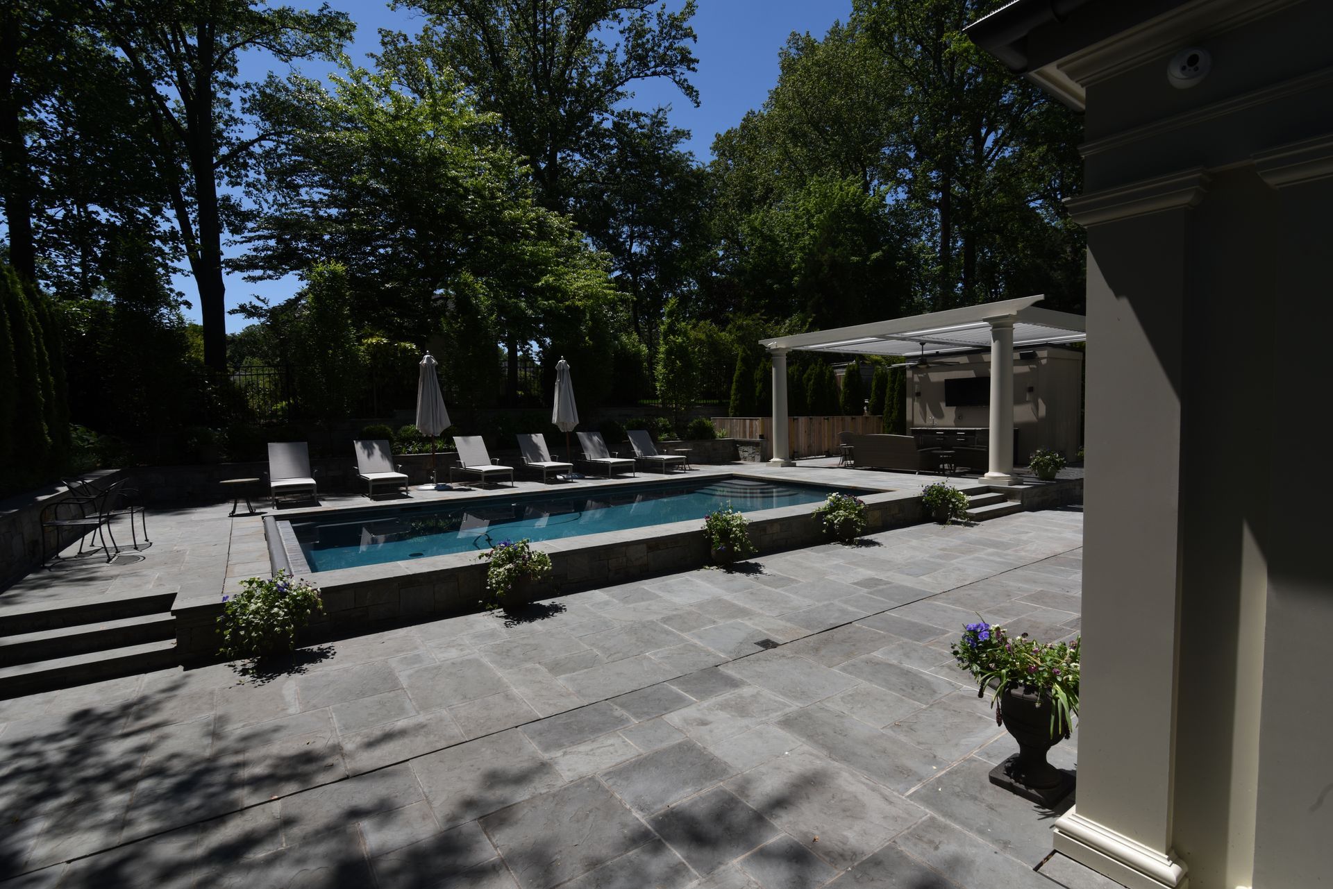 A rectangular pool with lounge chairs on a stone patio, surrounded by trees and a shaded pergola area.
