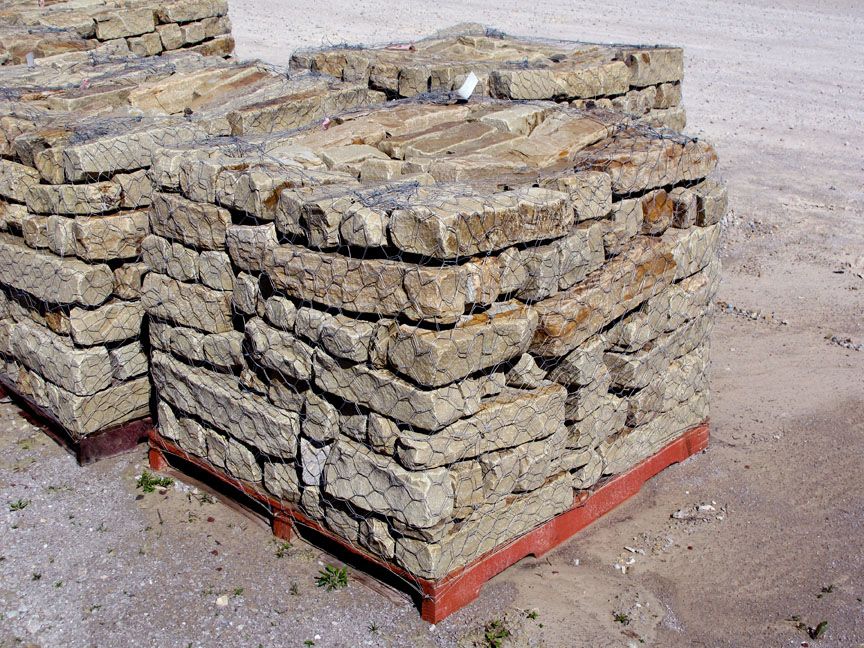 Two large, stacked square pallets of rough, light brown natural fieldstone on a dirt surface.
