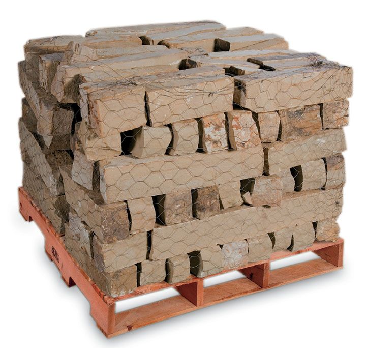 A pallet loaded with stacked, rustic tan stone blocks wrapped in thin wire mesh, isolated against a white background.