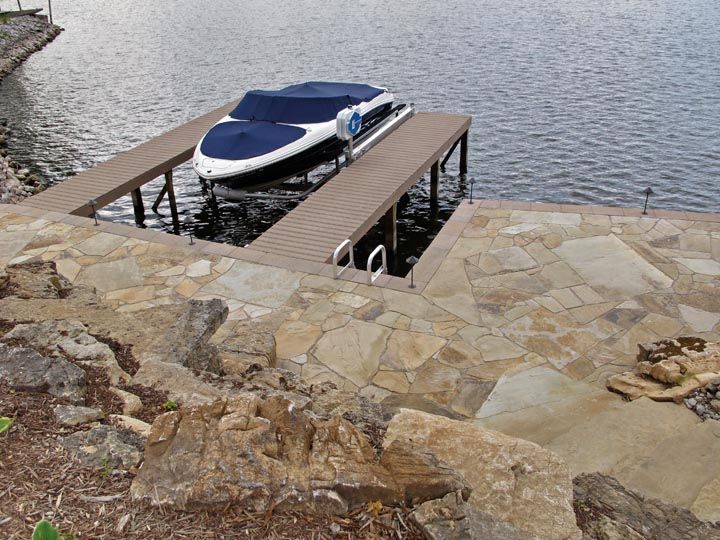 A boat with a blue cover is parked in a U-shaped dock next to a stone-paved patio by the water.