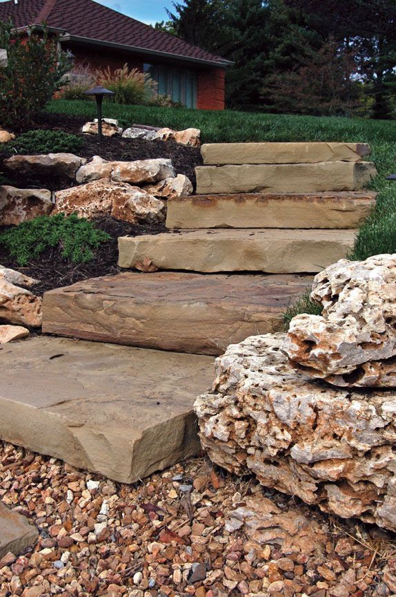 Stone steps lead up a grassy incline, flanked by large, textured landscape boulders and a gravel pathway.