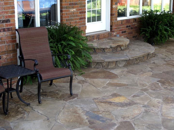 A brown outdoor chair and side table sit on a flagstone patio near a brick house with a stone step entry.